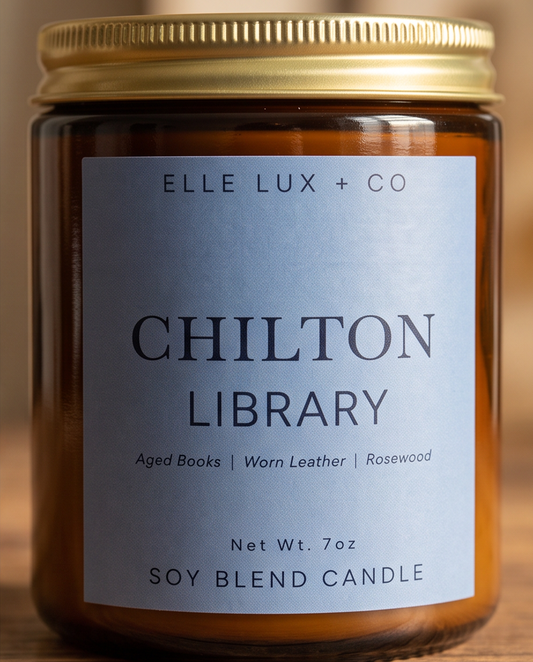 A close-up of an amber glass jar candle with a gold metal lid and a light blue label reading 'ELLE LUX + CO CHILTON LIBRARY' on a warm wooden background.