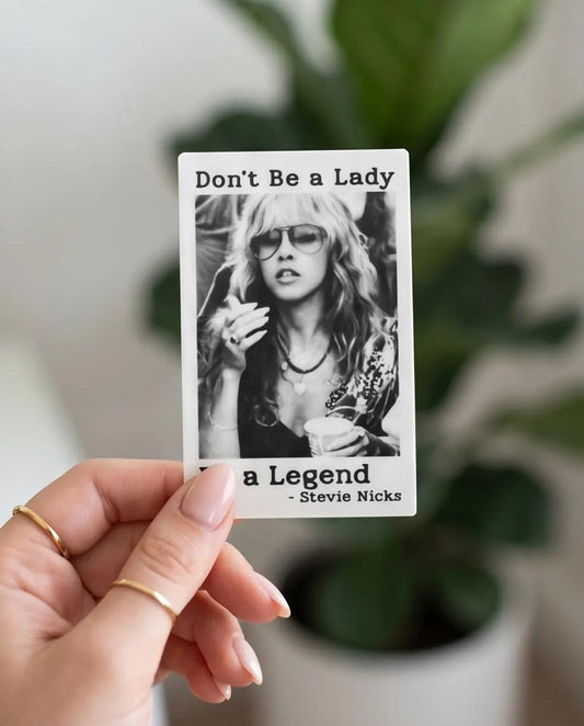 A hand holding a rectangular printed card that reads 'Don't Be a Lady - Stevie Nicks' with a black-and-white portrait photo in the center, against a softly blurred indoor background with a plant.