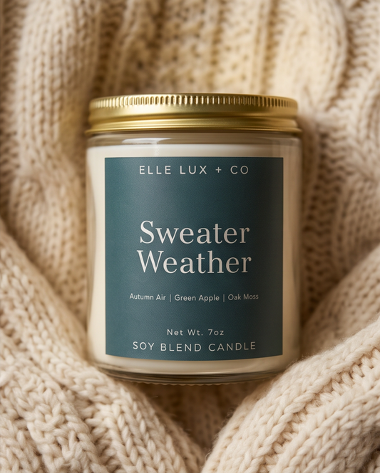 A 7oz soy blend candle by Elle Lux + Co in a clear glass jar with a gold lid and a slate blue label reading 'Sweater Weather'. The candle is styled on a cozy, cream-colored cable-knit sweater.