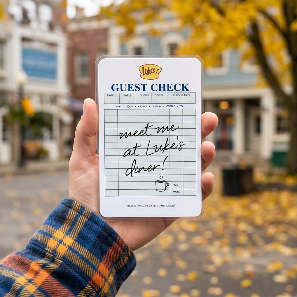 Hand holding a Gilmore Girls inspired Luke's Diner guest check bookmark in front of a cozy autumn streetscape reminiscent of Stars Hollow, perfect for book lovers and coffee fans.